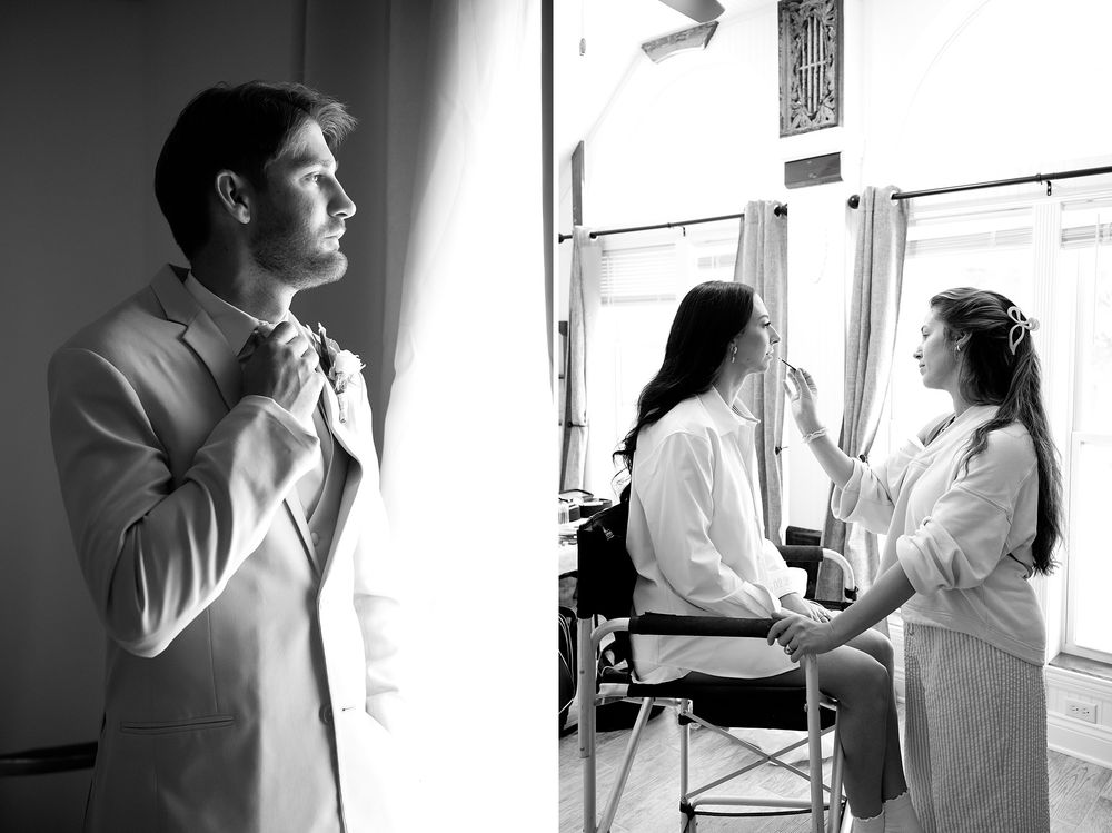 Black and white photo of groom adjusting tie while bridesmaids in robes prepare in background.