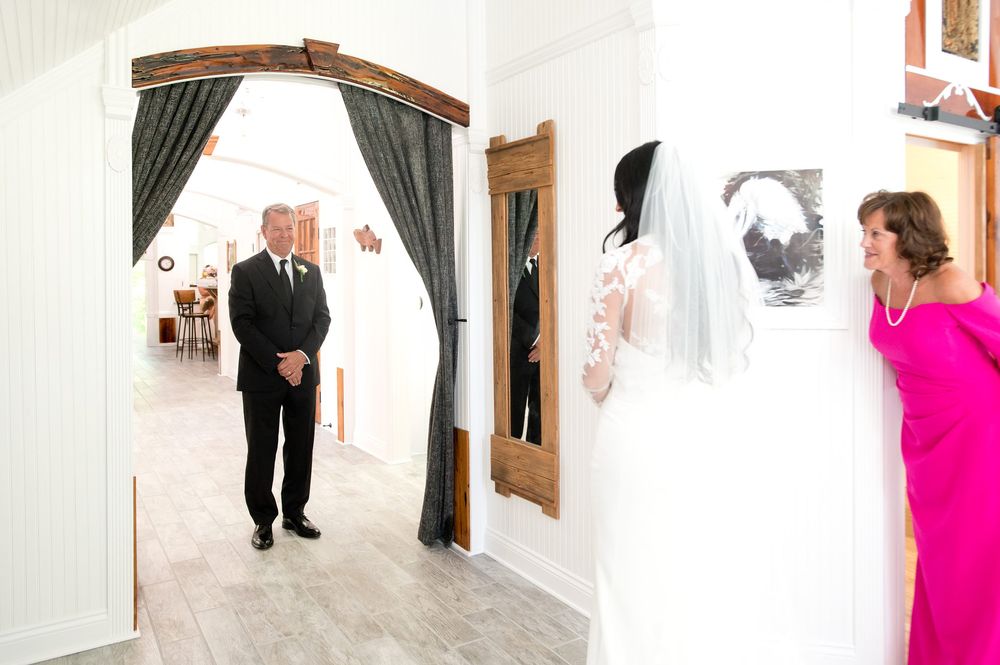Bride in white gown and veil stands in hallway as man in suit and woman in pink dress look on admiringly.