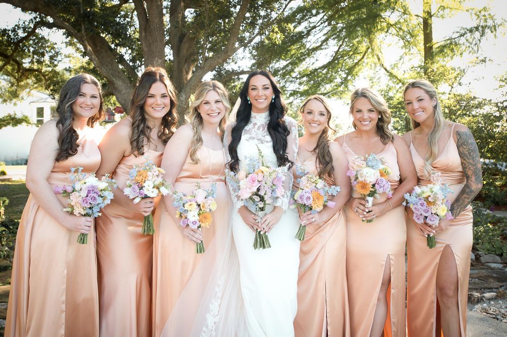 Bride in white lace gown poses with six bridesmaids in peach satin dresses holding colorful bouquets outdoors.