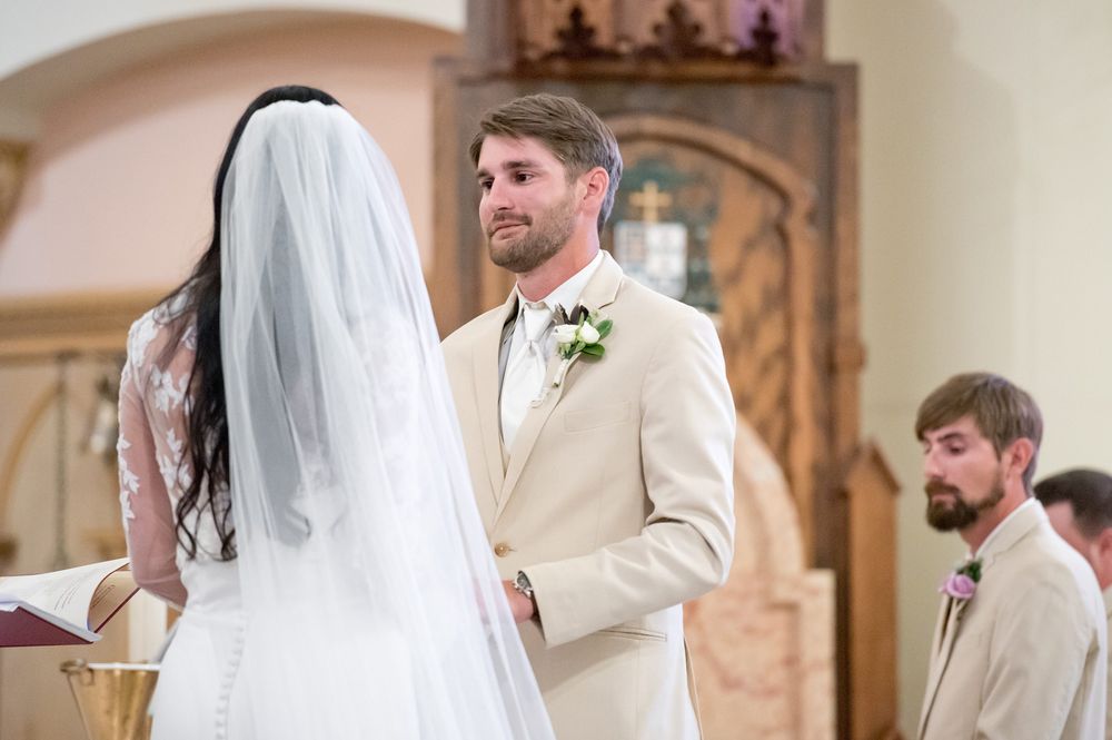 Groom in beige suit exchanges vows with bride in white veil at church wedding ceremony altar.