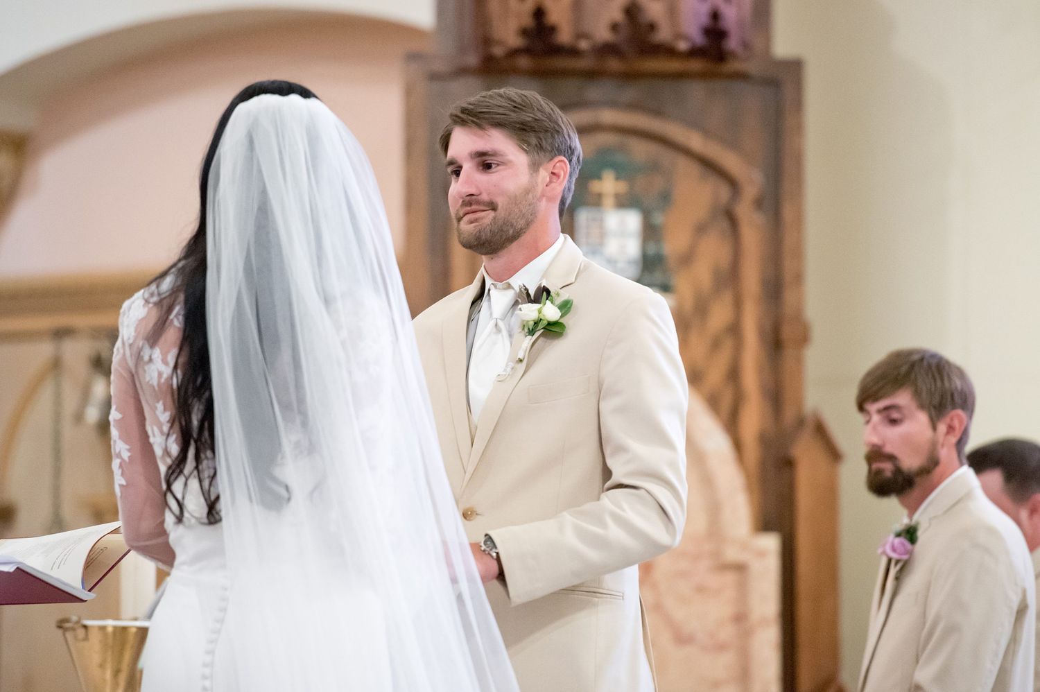 Groom in beige suit exchanges vows with bride in white veil at church wedding ceremony altar.