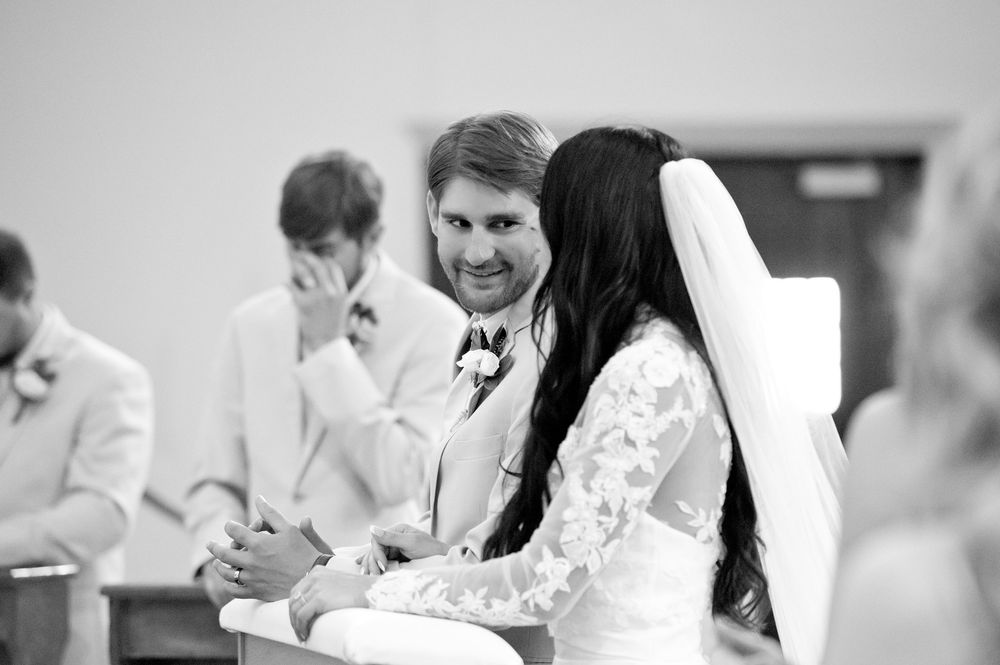 Black and white wedding photo of smiling groom gazing at bride in veil during ceremony.