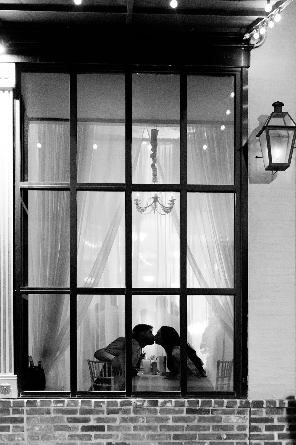 Black and white photo of a couple kissing silhouetted behind large glass windows with elegant curtains and chandelier.