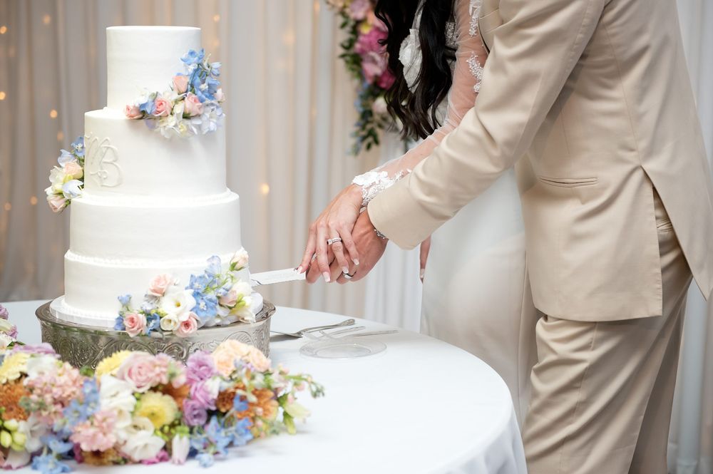 Bride and groom cutting a white four-tier wedding cake adorned with colorful flowers at their reception.