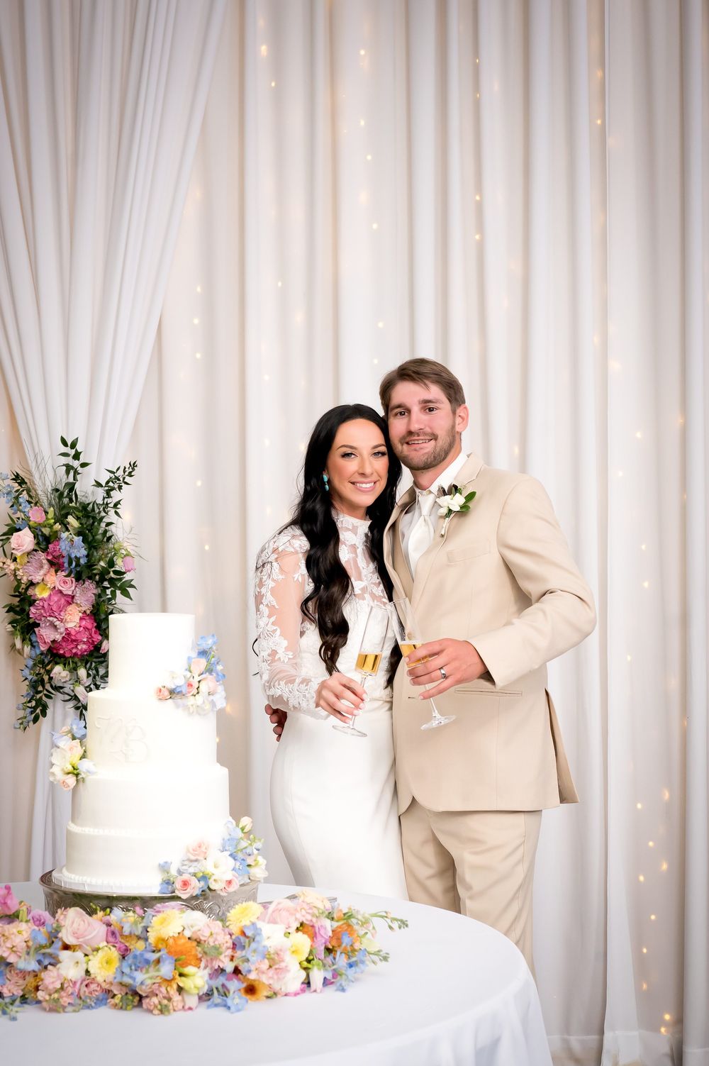 Bride and groom smiling beside a white wedding cake with champagne glasses and colorful floral arrangements.