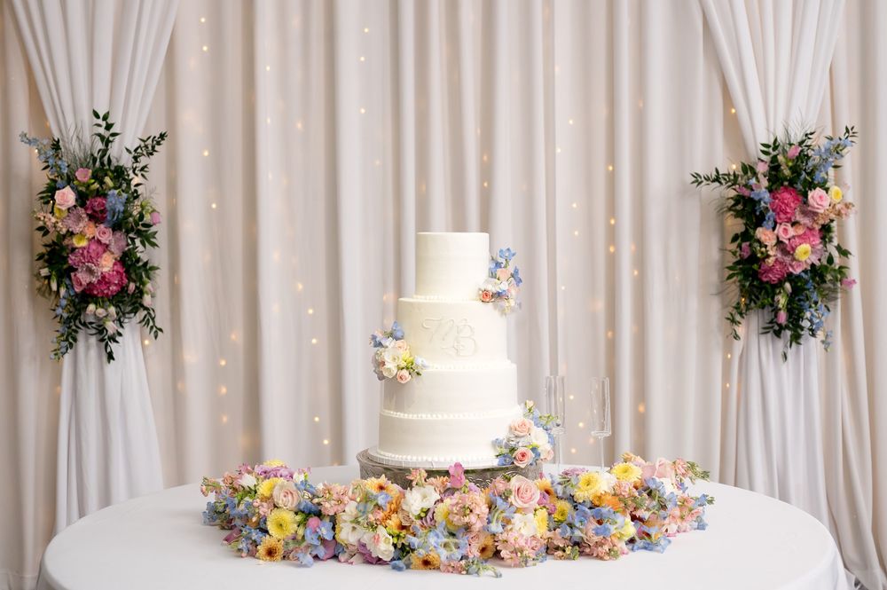 Elegant four-tier white wedding cake adorned with pastel flowers on a table with floral garland against draped backdrop.