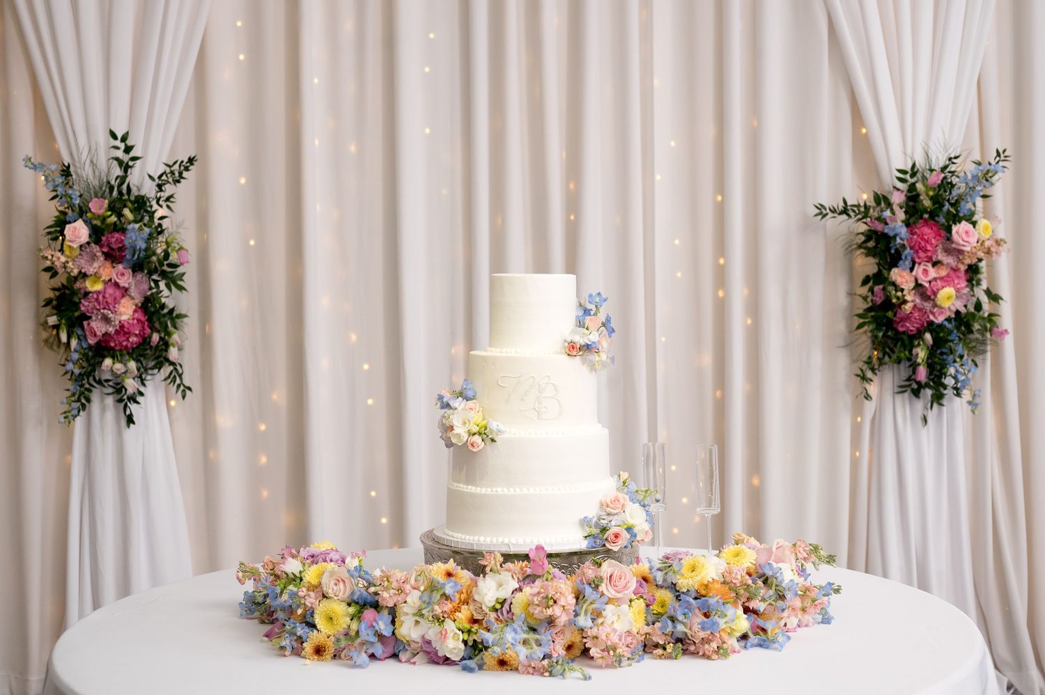 Elegant four-tier white wedding cake adorned with pastel flowers on a table with floral garland against draped backdrop.