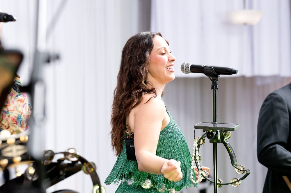Woman in green fringe dress performing on stage at an outdoor event, singing into a microphone.