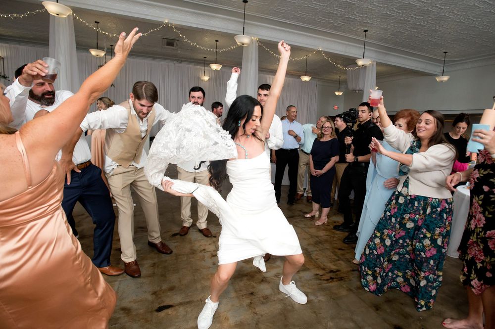 Bride in white dress and sneakers dancing energetically at wedding reception surrounded by guests celebrating on dance floor.
