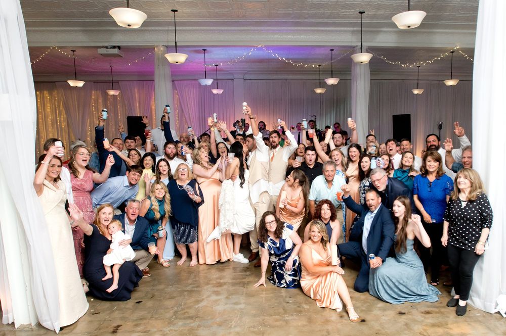 Large wedding party group photo with bride and groom surrounded by smiling guests in elegant venue.