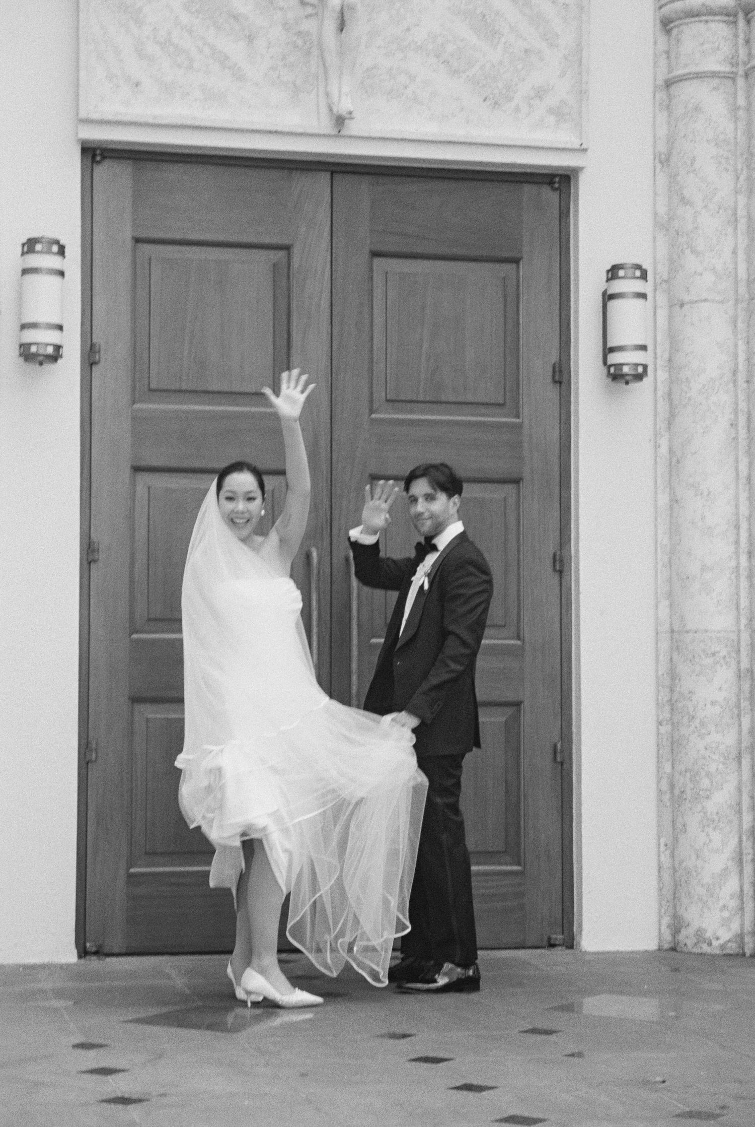Black and white photo of newlyweds waving joyfully outside church wooden doors after ceremony.