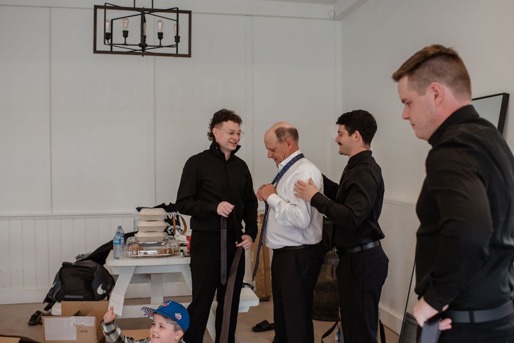 Wedding party members in black suits interact in a minimalist white room setting.