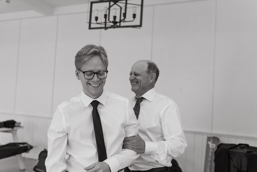 Groomsmen in white shirts and black ties sharing laughs in a white-walled room.