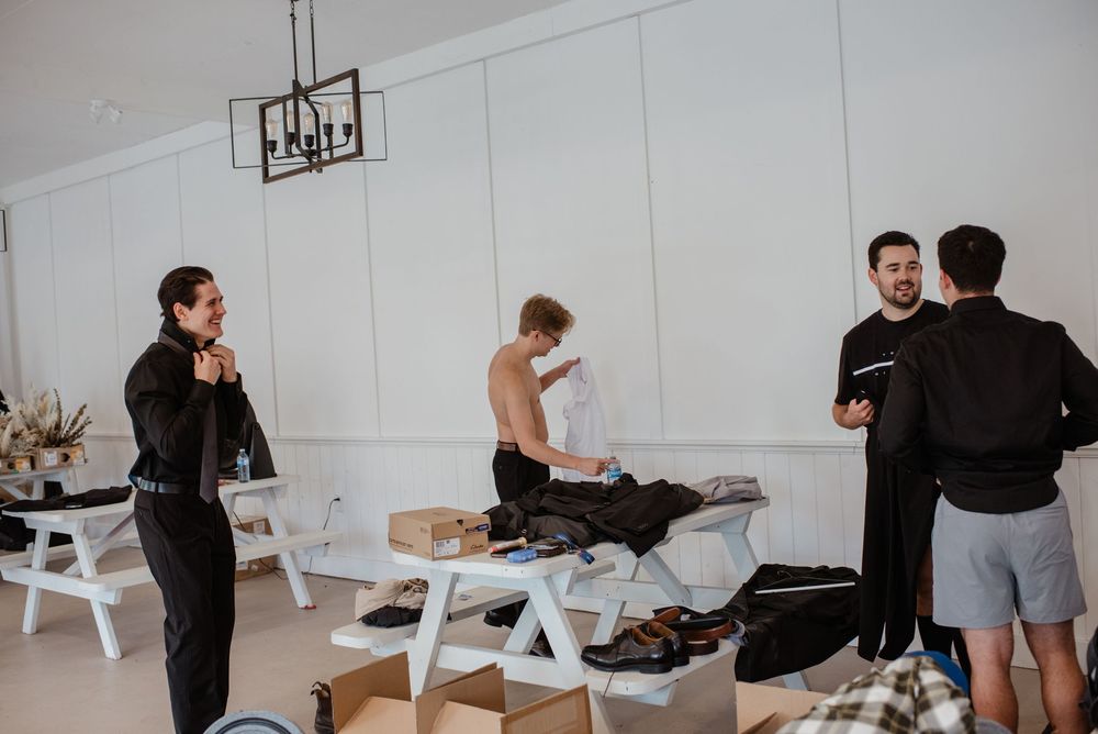 Men in formal attire gather around a table in a bright interior space during a wedding preparation event.
