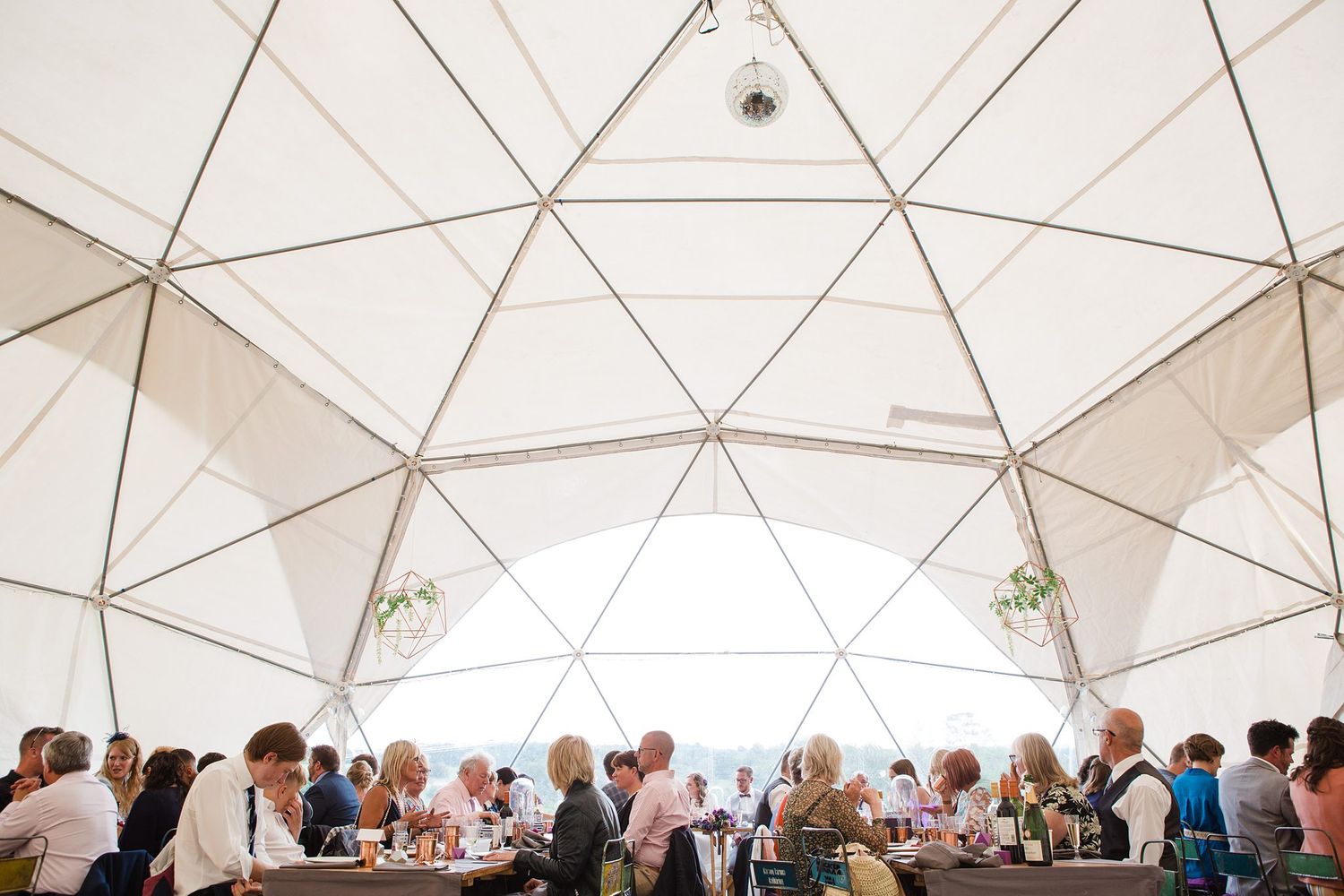 Guests dining under a geometric dome tent with white panels and wooden frame at an outdoor event.