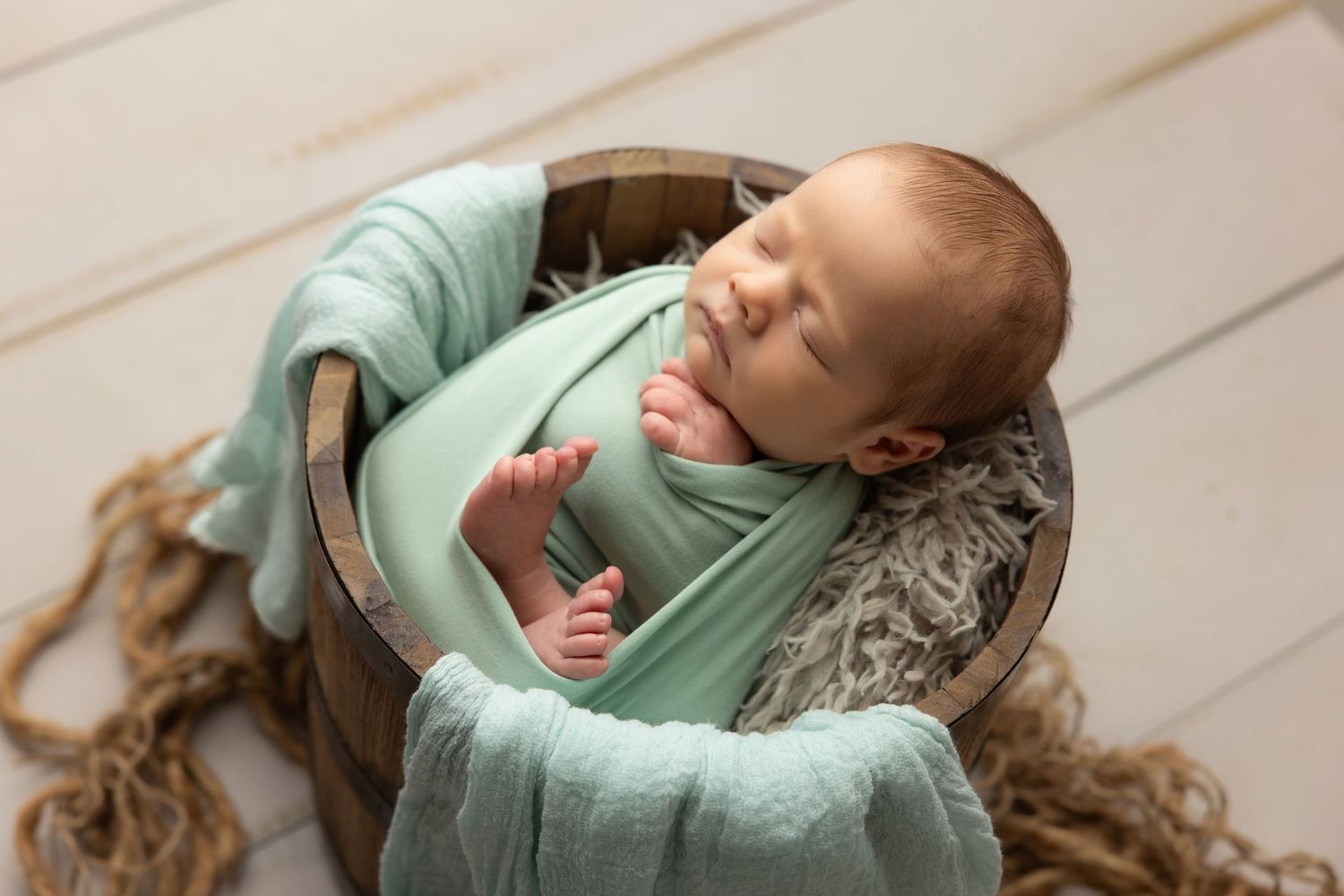 Newborn baby in teal hooded knit onesie with wooden buttons, sitting in a vintage metal bucket with soft teal blankets.