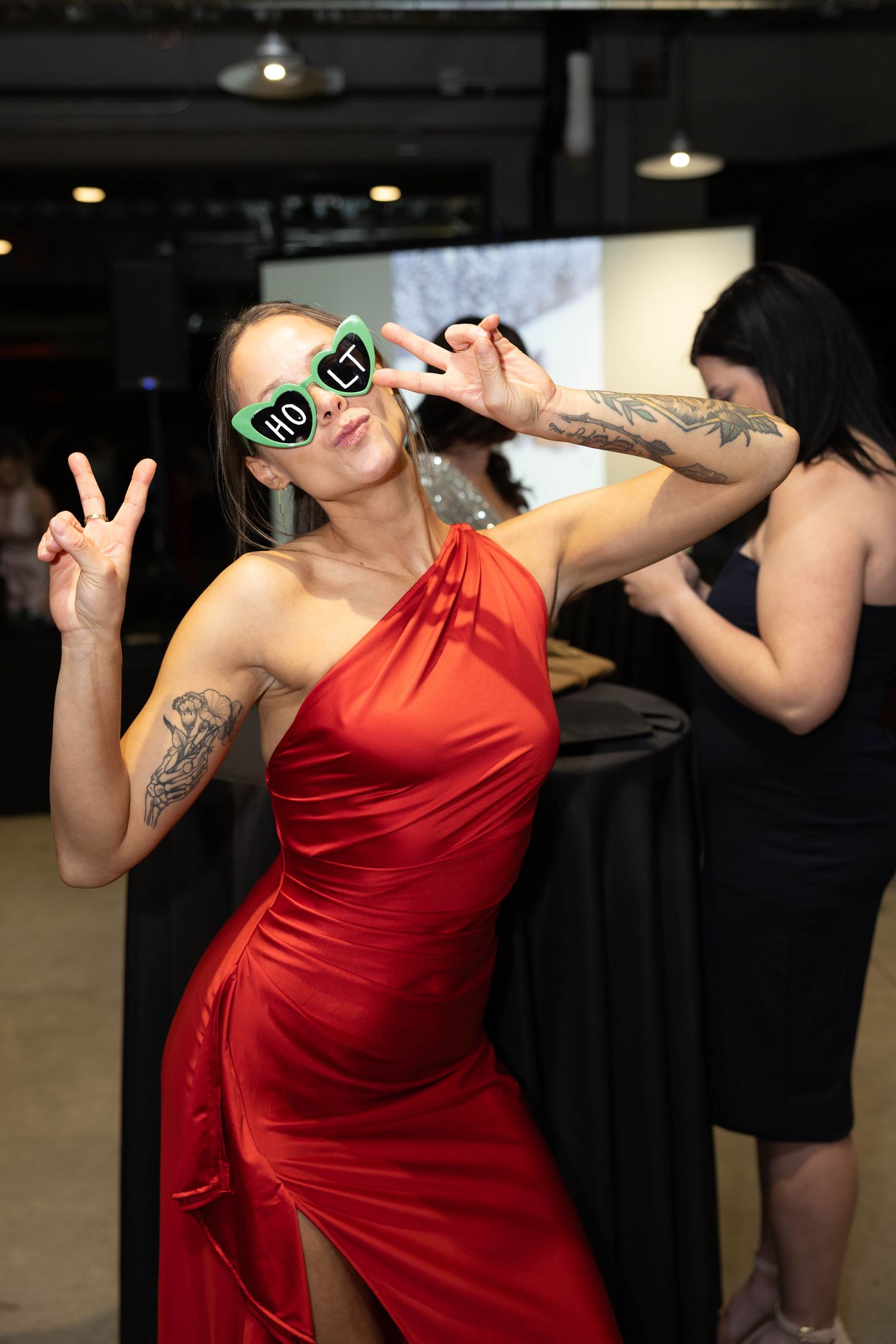 Woman in red dress with novelty sunglasses making peace signs at an indoor event.