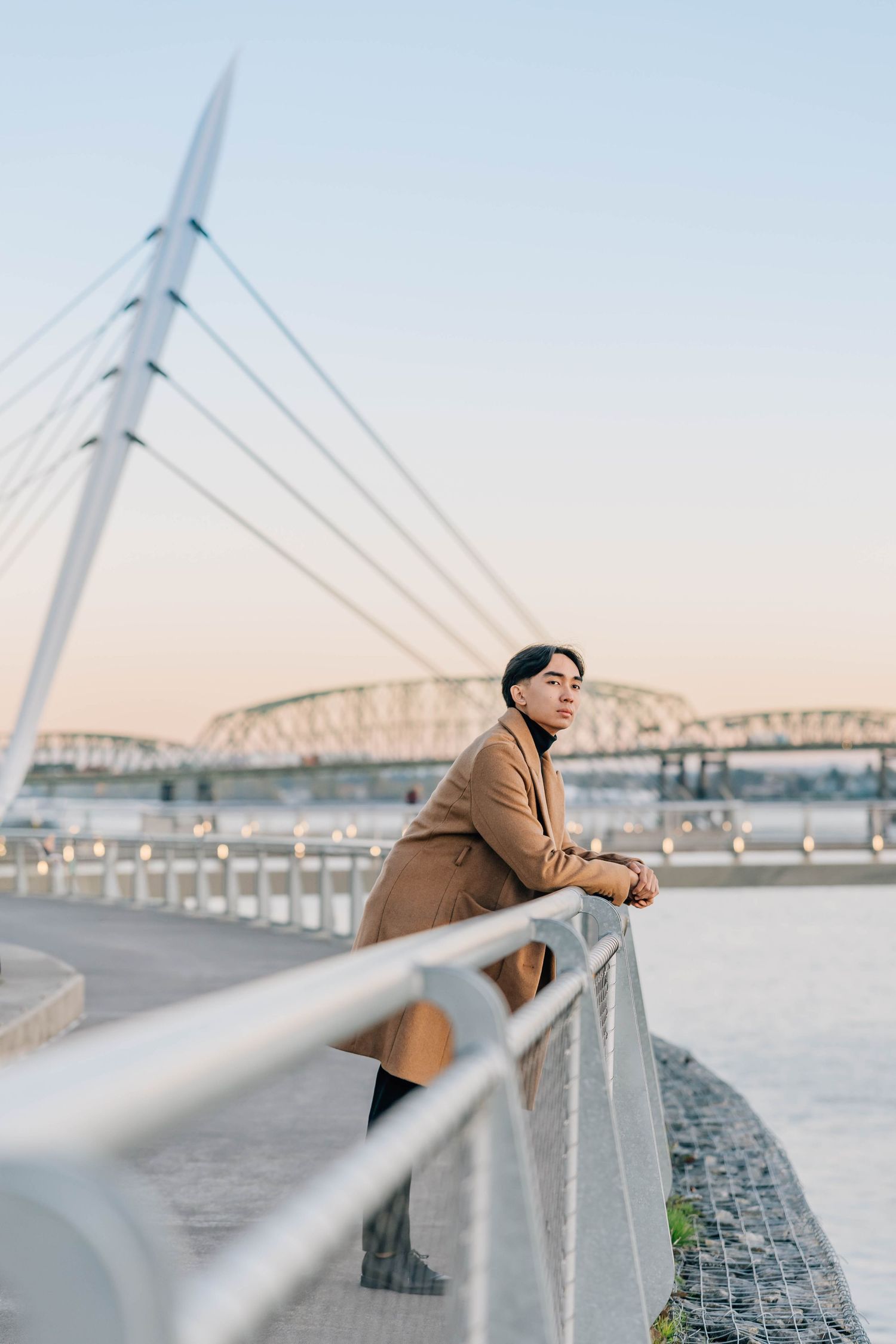Young man in beige coat leaning on bridge railing at sunset with cable bridge in background.