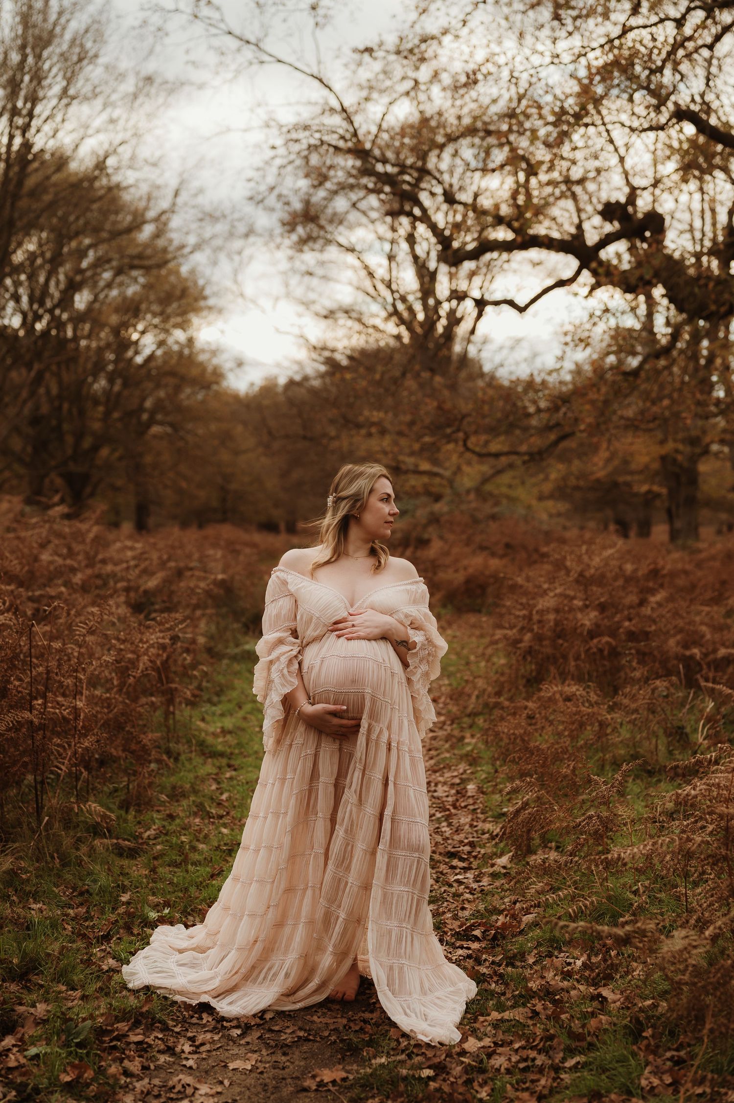 Pregnant woman in flowing blush gown stands among autumn ferns and bare trees in a serene woodland maternity portrait.