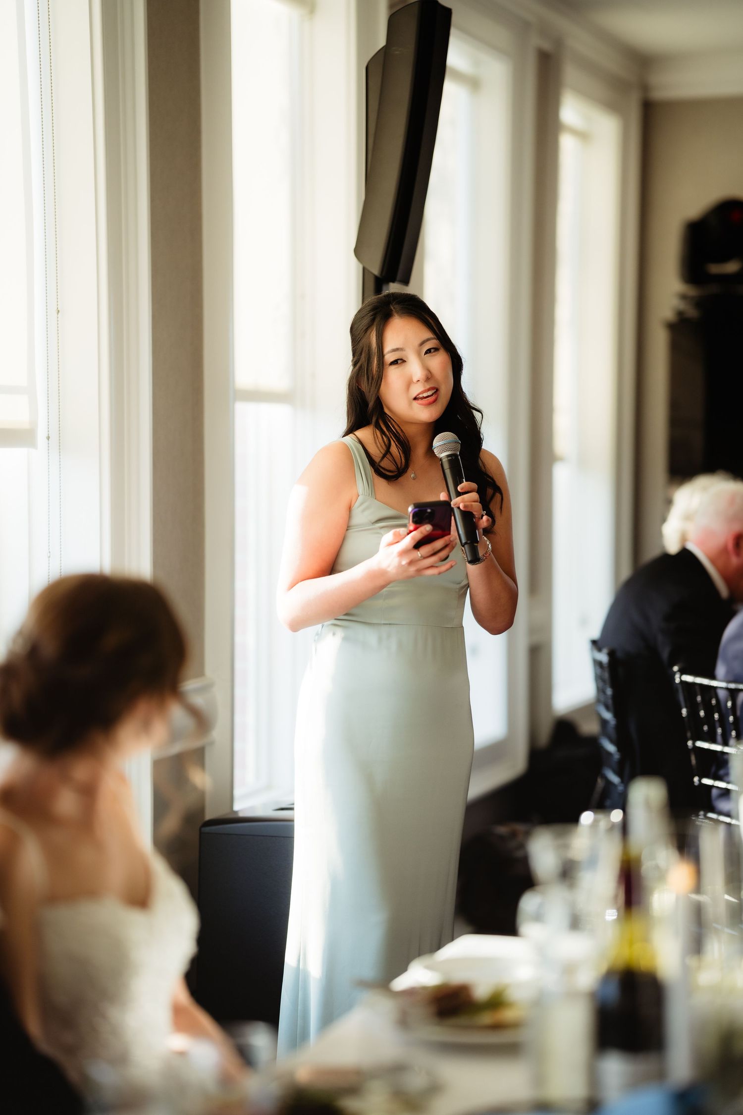 Bridesmaid in mint green gown giving a speech with microphone at an elegant wedding reception.