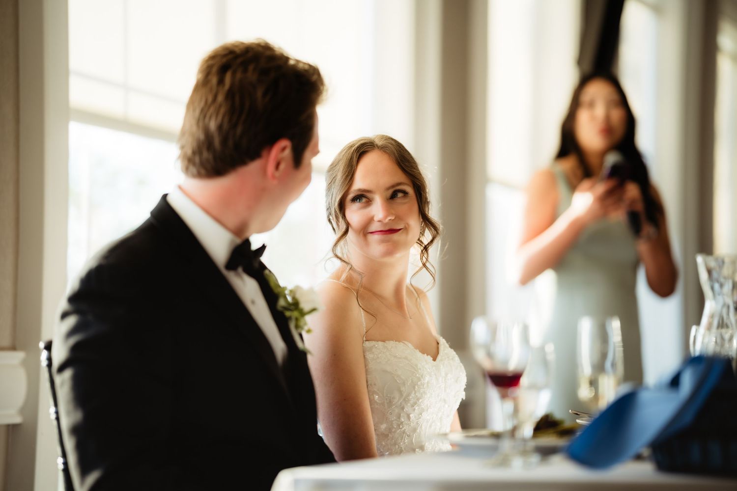 Bride smiles lovingly at groom in tuxedo during wedding reception, with bridesmaid photographing in background.