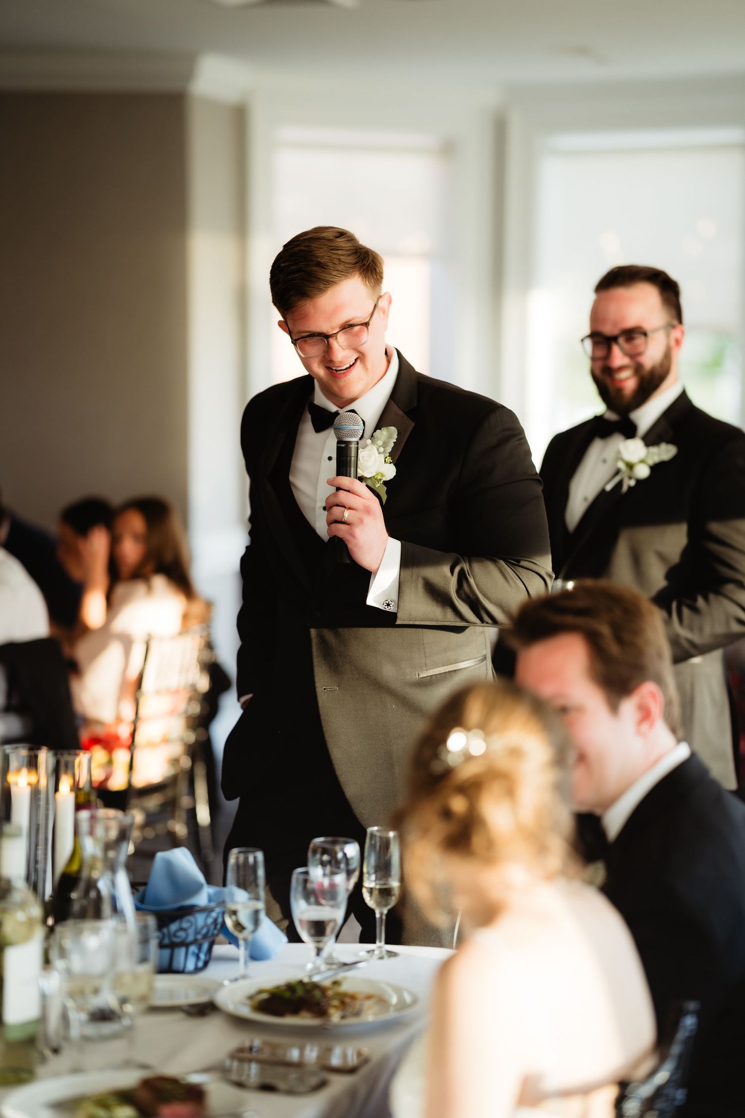 A smiling groomsman delivers a wedding toast with a microphone at an elegant reception dinner.