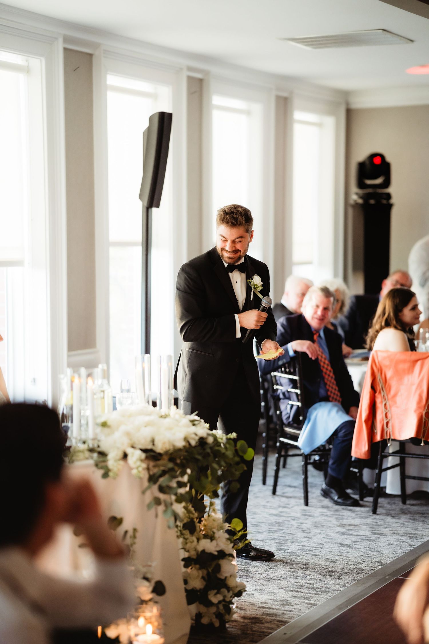 Groom in tuxedo giving speech at elegant wedding reception with floral arrangements and seated guests.