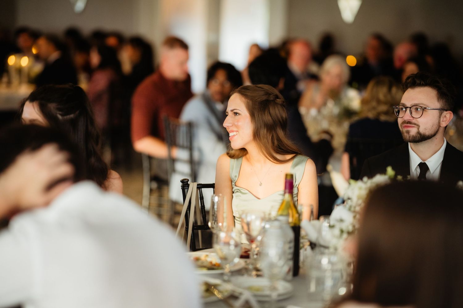 Guests smiling and socializing at an elegant wedding reception dinner, seated at decorated tables with wine bottles.
