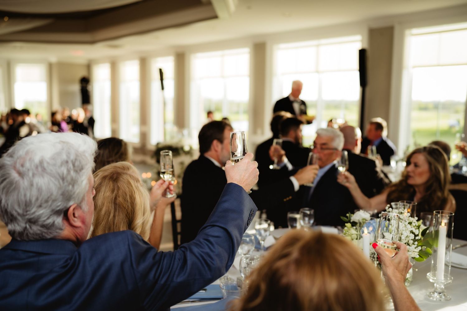 Guests raising champagne glasses in a toast at an elegant wedding reception dinner in a bright venue.
