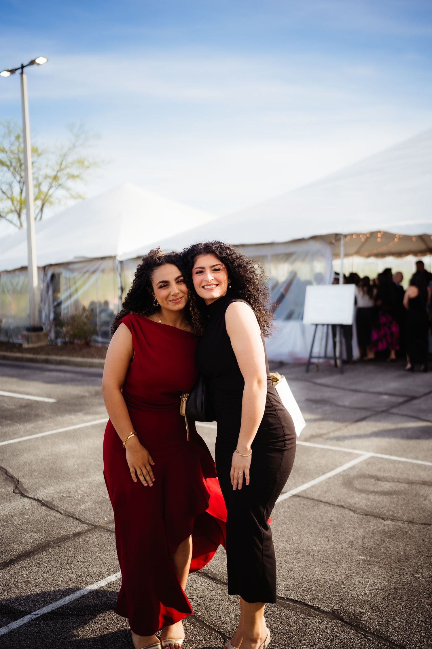 Two women in elegant red and black outfits posing together outdoors near a white event tent.