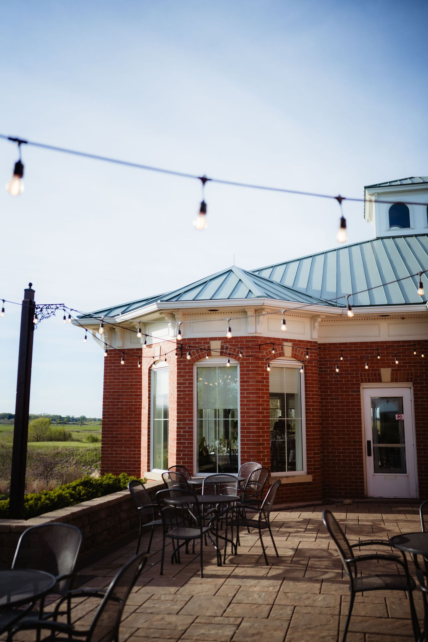 Brick building patio with string lights, metal chairs, and tables under a blue sky at dusk.