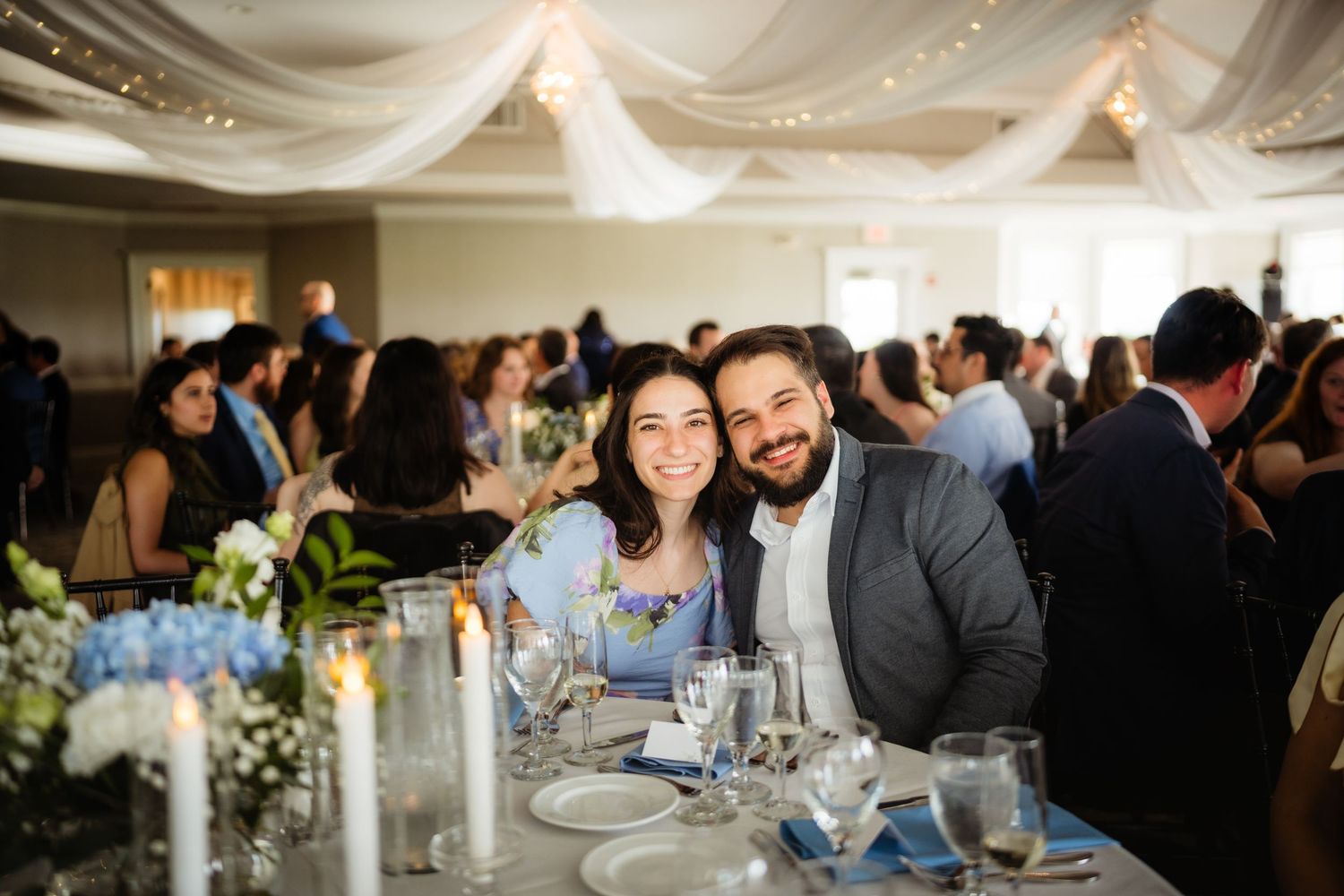 Smiling couple poses together at an elegant wedding reception with draped ceiling and floral centerpieces.