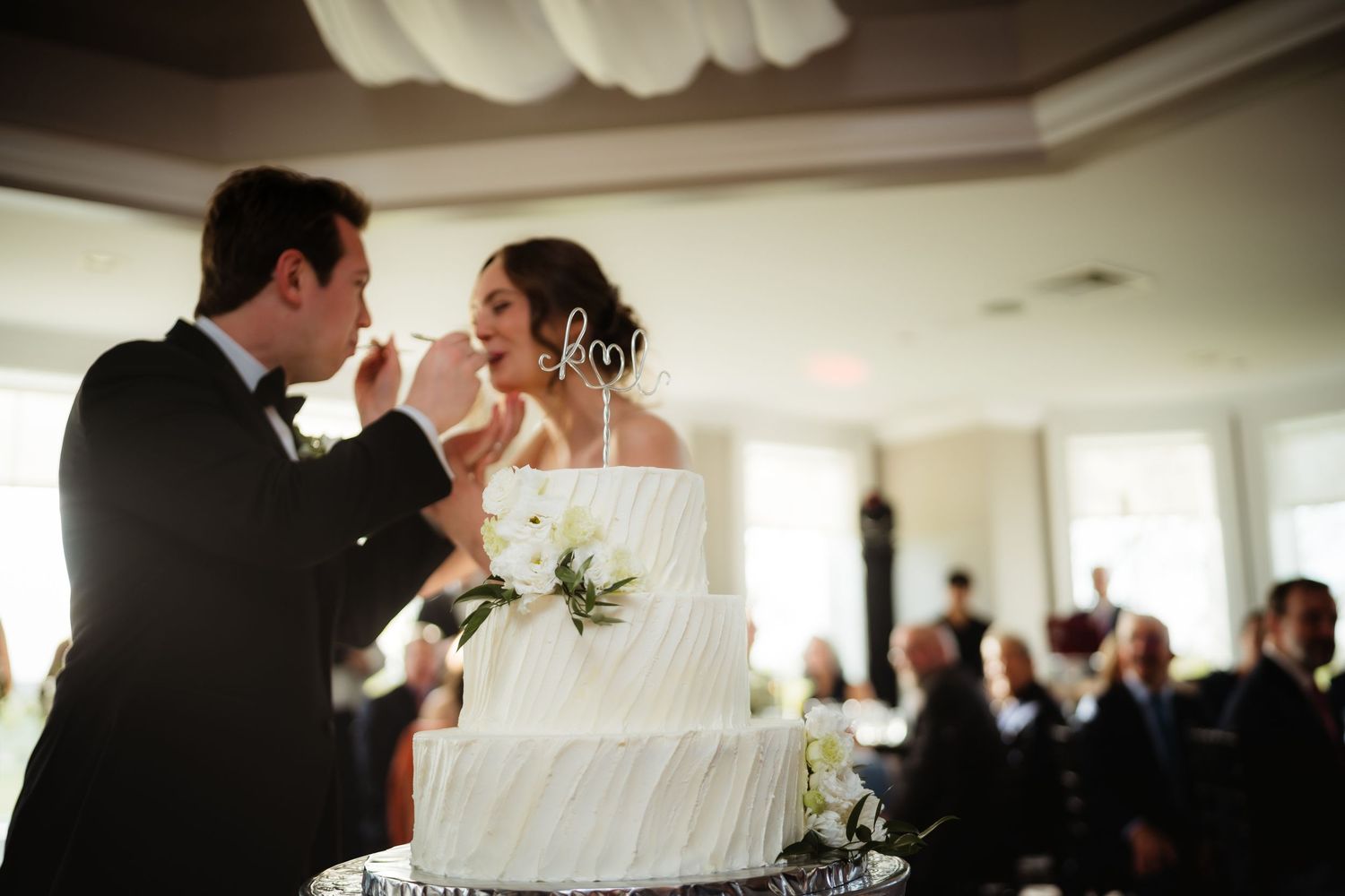Bride and groom cutting and sharing their elegant white tiered wedding cake at reception.