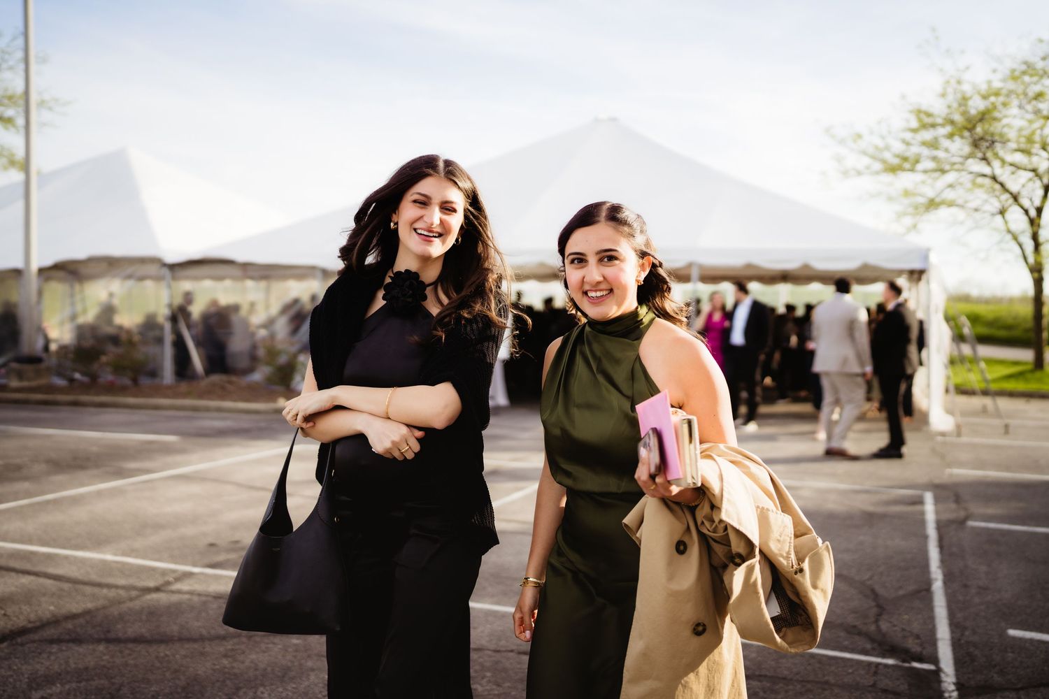 Two smiling women in elegant attire pose outdoors near a white event tent at a formal gathering.