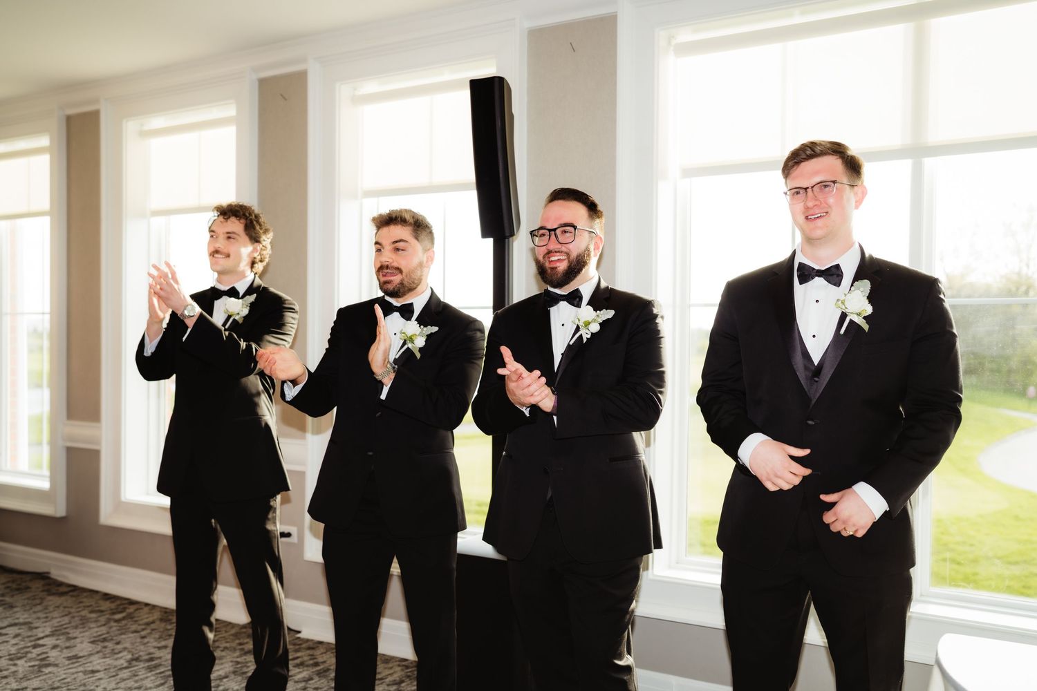 Four groomsmen in black tuxedos with white boutonnieres clapping and smiling at a wedding reception.
