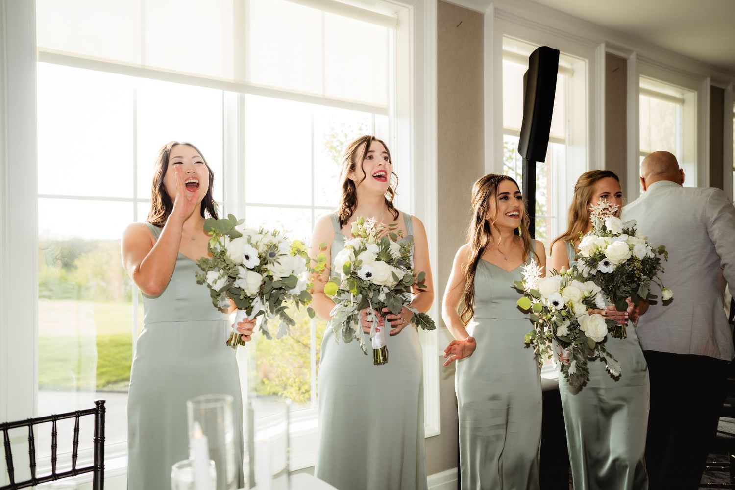 Three bridesmaids in sage green gowns holding white floral bouquets react with joy during a wedding ceremony indoors.