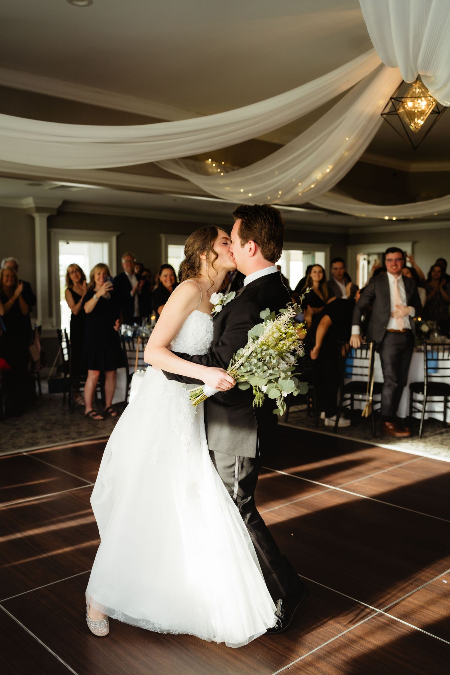Bride and groom share first dance kiss in decorated ballroom, guests watching as bride holds green bouquet.