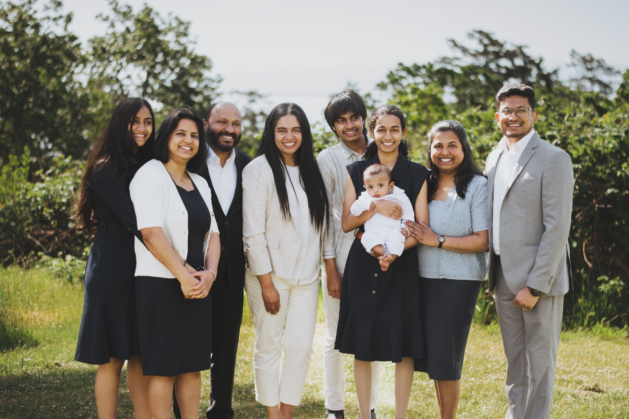 Large multigenerational South Asian family posing together outdoors, smiling, with lush green trees in the background.