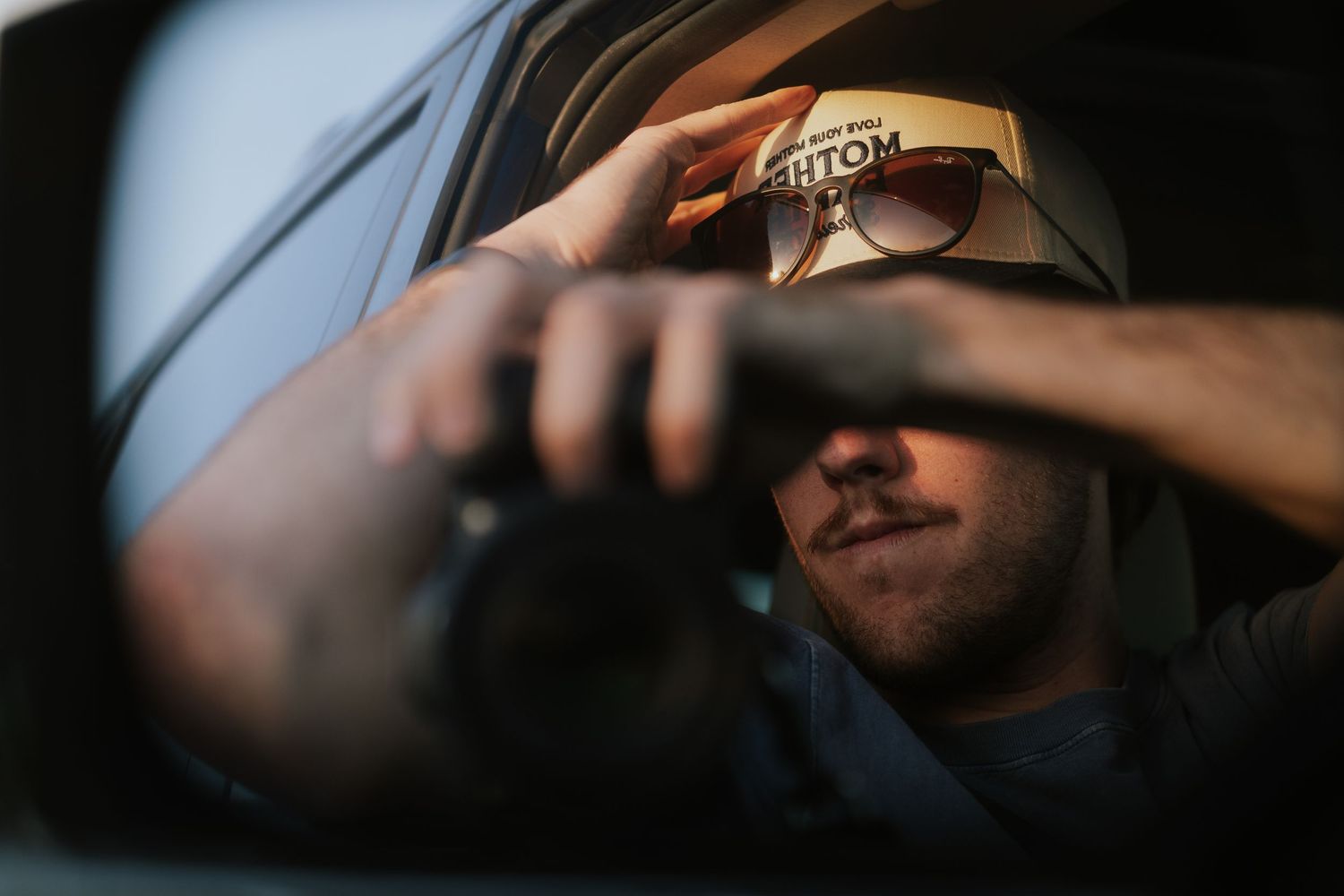 Man wearing sunglasses and cap photographing with camera, reflected in car side mirror.