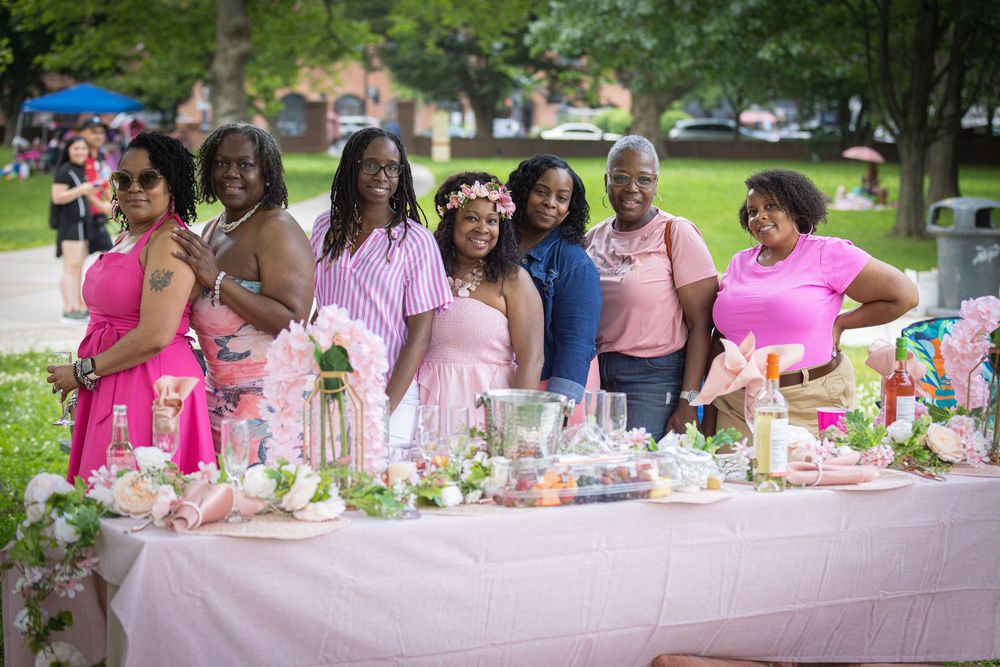 Seven women in pink attire smile together at an outdoor party table decorated with flowers and elegant pink decor.