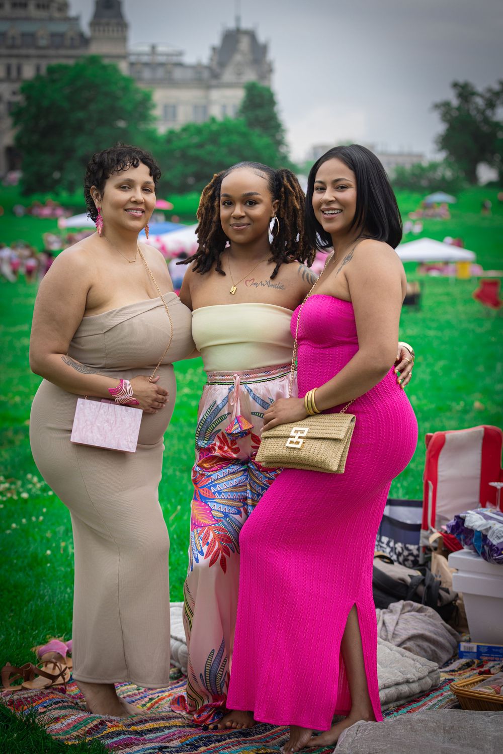 Three stylish women posing outdoors at a park event, wearing elegant outfits in beige, colorful, and pink dresses.
