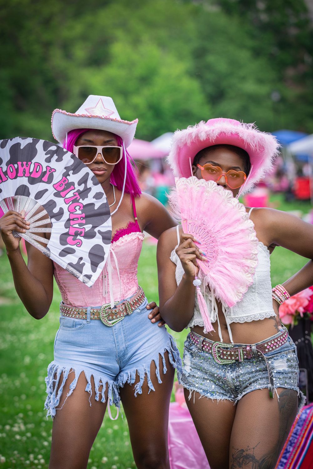 Two women in pink cowboy hats, denim shorts, and corsets pose with decorative fans at an outdoor festival.