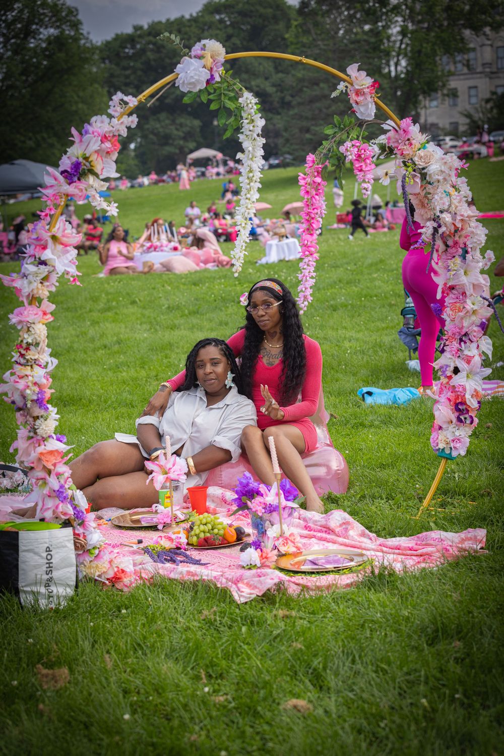 Two women sit together under a floral arch at a pink-themed outdoor picnic on a green lawn.
