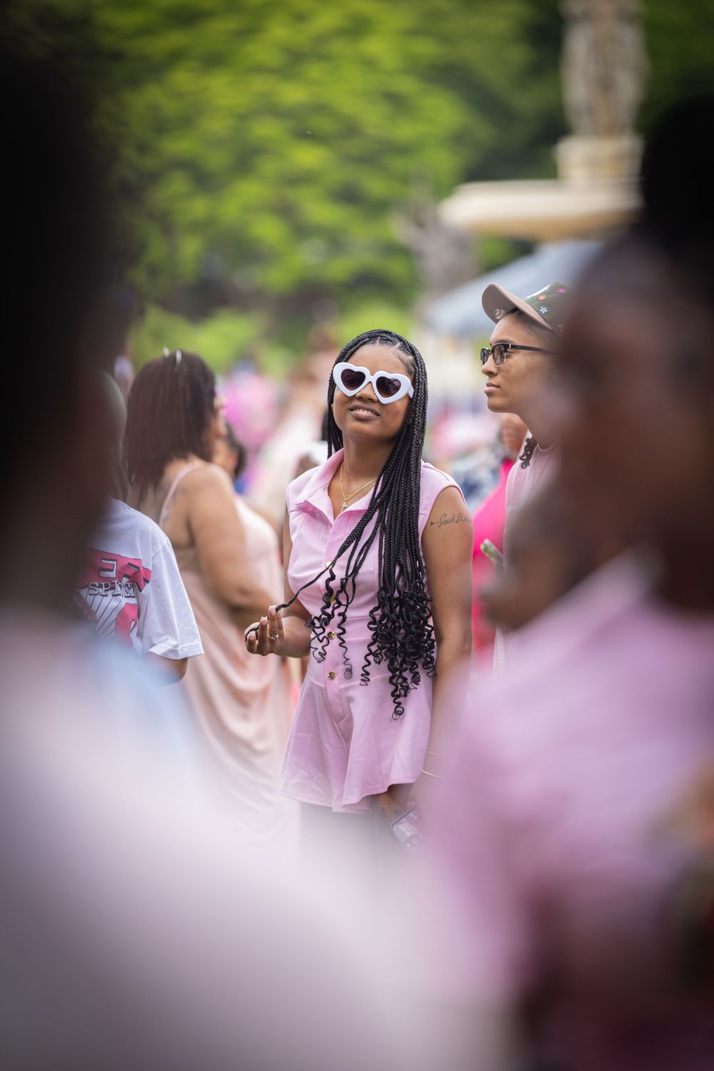 Young Black woman in pink dress and heart-shaped white sunglasses stands out in a crowd at an outdoor event.