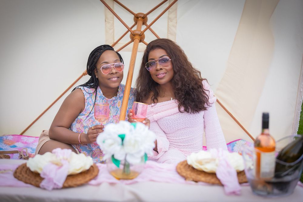 Two women smiling and holding wine glasses at a decorated picnic table under a canopy tent.