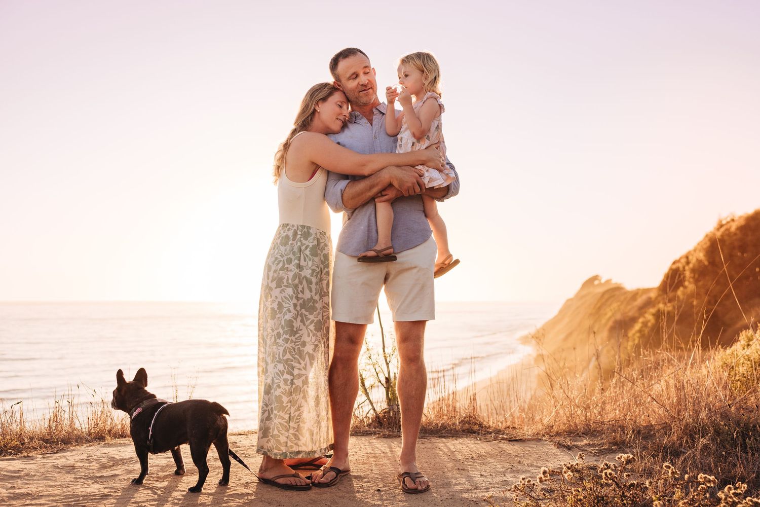 Happy family with toddler and French dog posing on coastal cliff at golden sunset what to wear for bluffs photo session