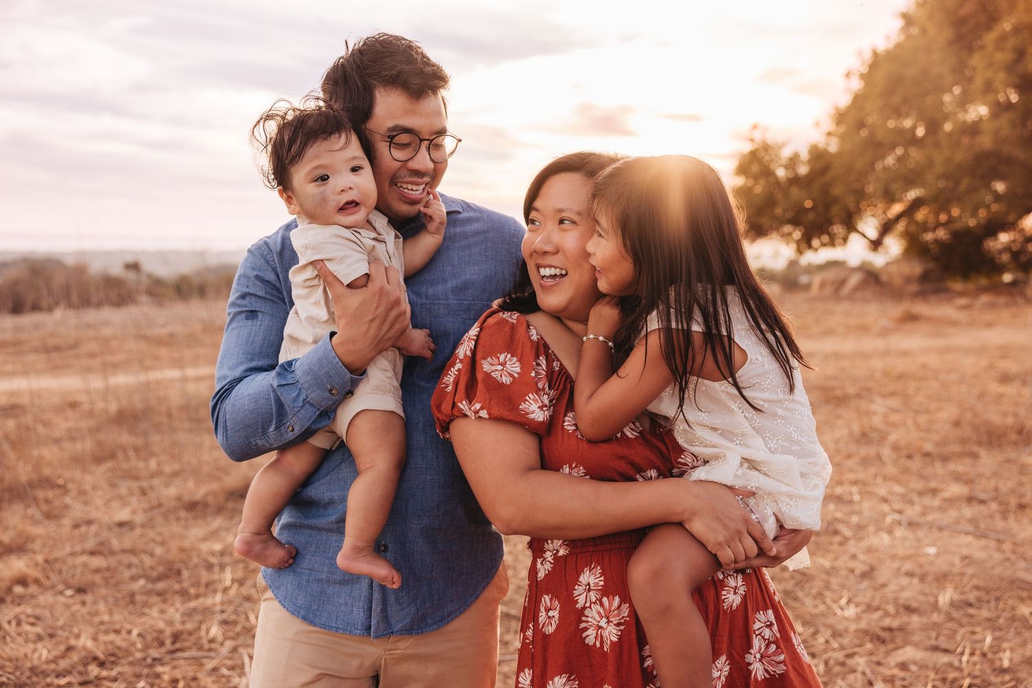 Family of four walking on a sunny dirt path surrounded by golden fields and trees during a warm sunset.