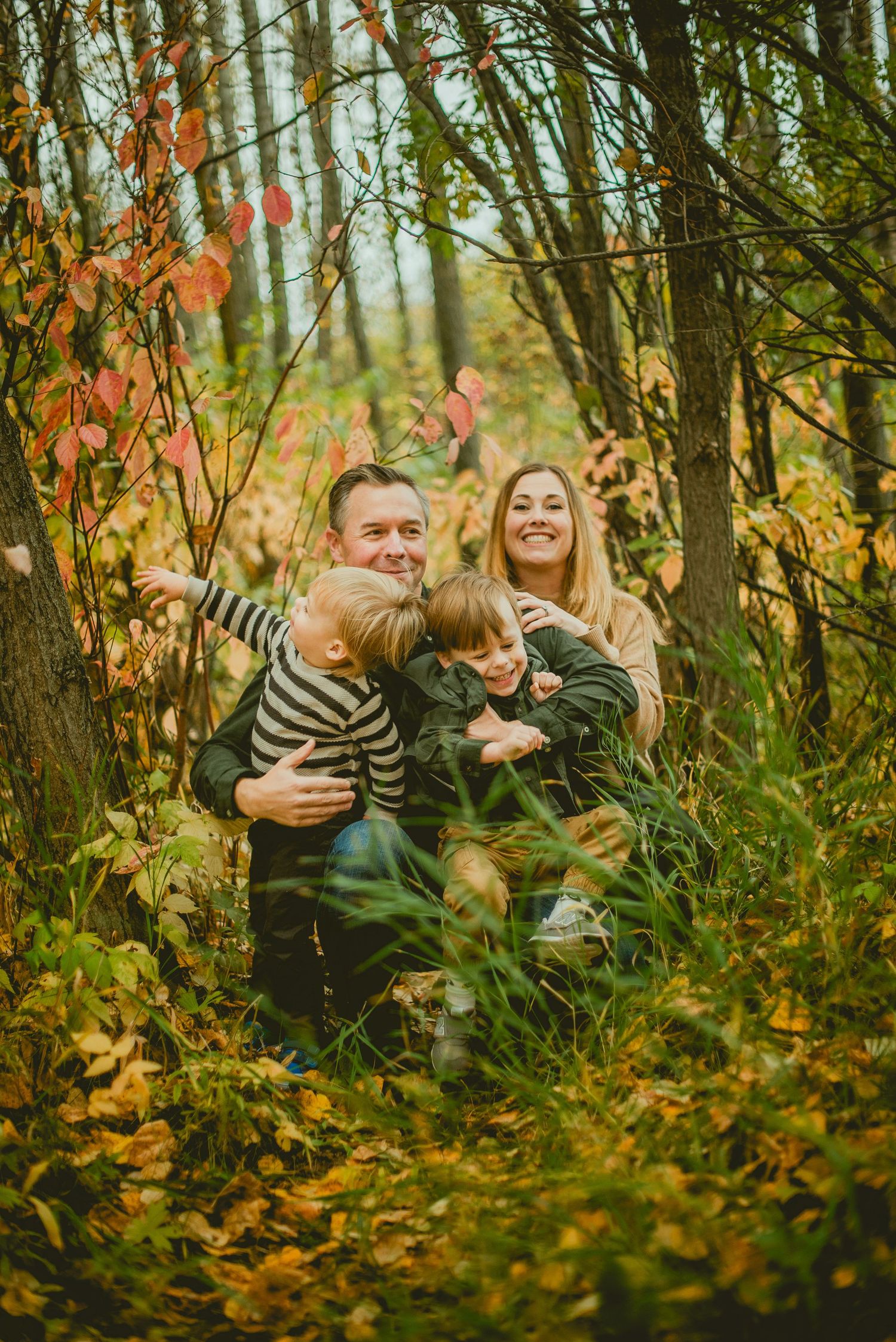 Family of four laughing together in an autumn forest surrounded by colorful fall foliage.