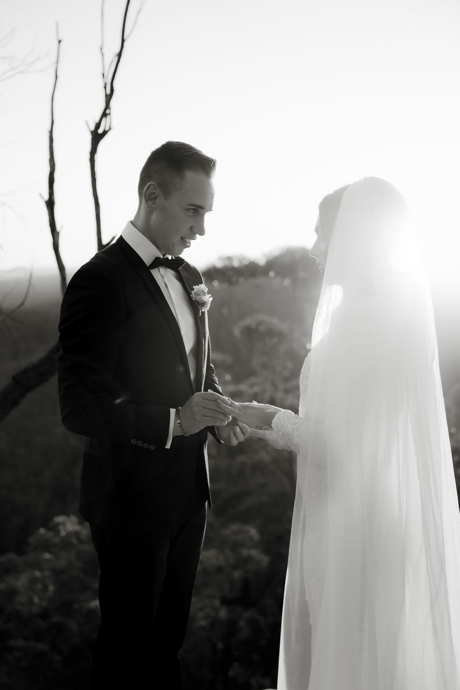 Black and white photo of groom placing wedding ring on brides hand during outdoor ceremony with soft backlight.