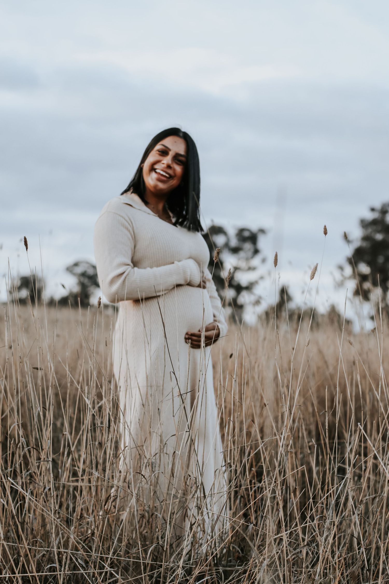 Smiling pregnant woman in white dress standing in golden grass field, cradling baby bump outdoors.